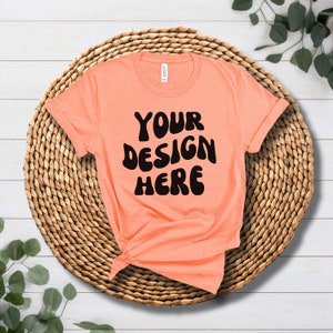 May include: A peach coloured t-shirt with the text "YOUR DESIGN HERE" in black, on a woven straw mat.