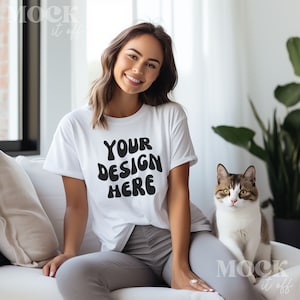 Pode incluir: Uma mulher vestindo uma camiseta branca com o texto "YOUR DESIGN HERE" em preto, sentada em um sofá branco com um gato ao seu lado.