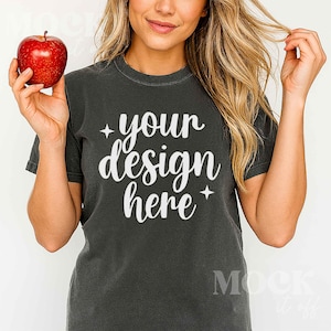 May include: Dark gray t-shirt with the text "your design here" in white script. A red apple is held in the left hand. The shirt has short sleeves and a crew neck. The image is a mock-up.
