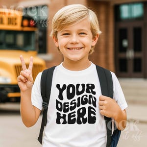 May include: A white t-shirt with the text "YOUR DESIGN HERE" in black, worn by a smiling child with a backpack. The child is giving a peace sign. A yellow school bus is visible in the background.