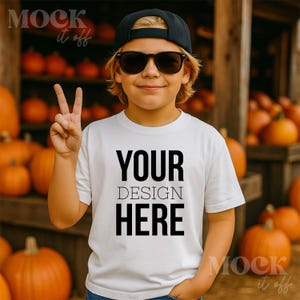 Op de afbeelding: Een wit t-shirt met de tekst "YOUR DESIGN HERE" in het zwart. De persoon draagt een zwarte pet en zonnebril en geeft het vredesteken. Pompoenen staan op de achtergrond.