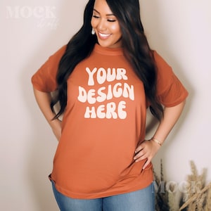 Puede incluir: Camiseta color óxido con el texto "YOUR DESIGN HERE" en una fuente retro. La camiseta de manga corta tiene cuello alto y un corte relajado. La modelo lleva vaqueros azules y una pulsera dorada.