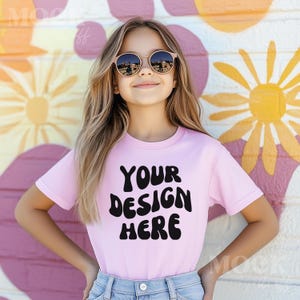 Puede incluir: Una persona joven lleva una camiseta rosa con el texto "YOUR DESIGN HERE" en letras negras de estilo retro. La persona lleva gafas de sol y está de pie frente a una pared colorida con un patrón de flores.