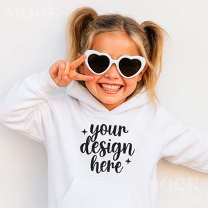 May include: A white hoodie with a front pocket and the text "your design here" in black script. The person is wearing white heart-shaped sunglasses with dark lenses. The person is smiling and giving a peace sign.