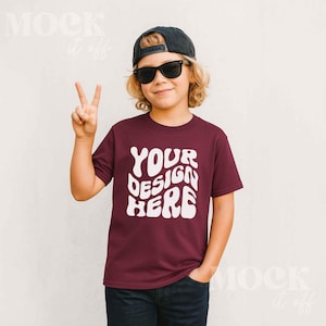 May include: A maroon t-shirt with the text "YOUR DESIGN HERE" in white, worn by a person wearing a black cap and sunglasses. The person is giving a peace sign.