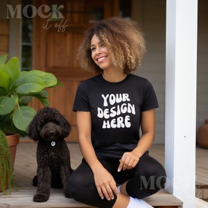 May include: A woman with curly hair is sitting on a porch with a black dog. She is wearing a black t-shirt with the text "YOUR DESIGN HERE" in white.