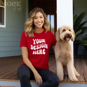 May include: A woman wearing a red t-shirt with the text "YOUR DESIGN HERE" in white. She is sitting on a wooden porch with a light brown dog.