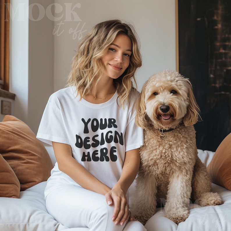 Pu&ograve; includere: Una donna indossa una maglietta bianca con il testo "YOUR DESIGN HERE" in nero, seduta su un divano bianco con un cane marrone chiaro.