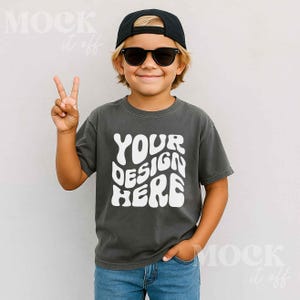May include: A child wearing a dark gray t-shirt with the text "YOUR DESIGN HERE" in white, a black baseball cap, and black sunglasses. The child is smiling and giving a peace sign.