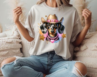 May include: A white t-shirt with a graphic of a dachshund wearing a pink and gold cowboy hat and a pink heart necklace. The dachshund is surrounded by gold glitter stars and a watercolor background.