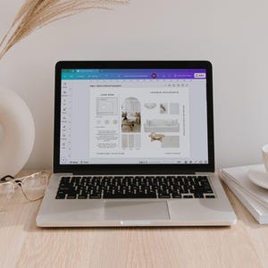 May include: A silver laptop displaying an interior design presentation on its screen, positioned next to a white donut-shaped vase with dried grass, clear-framed glasses, and a white coffee cup on a saucer resting on a stack of white books.