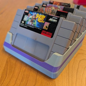 May include: A gray and purple 3D-printed video game cartridge holder filled with several Super Nintendo game cartridges. The front cartridge features the game "Scooby Doo Mystery". The holder is on a wooden surface.
