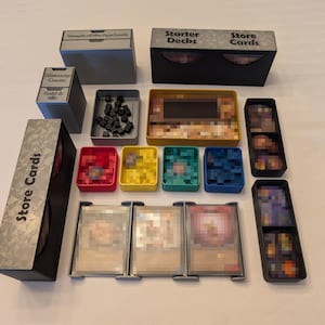 May include: A board game organizer set featuring black, gray, yellow, red, teal, and blue plastic containers. The set includes boxes labeled Starter Decks, Store Cards, and various trays for holding game cards, small black cubes, and tokens.