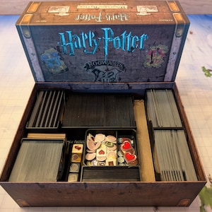 May include: A brown wooden box with a Harry Potter Hogwarts logo on the lid. The box is open and contains various game pieces, including cards, tokens, and dice.