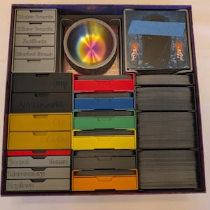 May include: A board game organizer insert featuring multiple colored plastic trays for cards and tokens. The gray trays are labeled Major Secrets, Minor Secrets, Artifacts, Market Items, Secret Tombs, Mercenary, and Explore. Includes a rainbow-colored bowl.