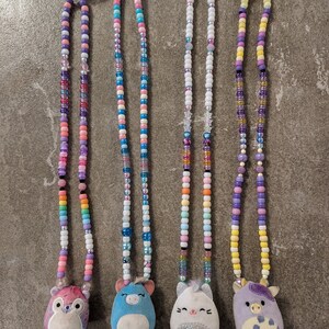 May include: Four beaded necklaces with plush animal pendants. The necklaces feature colorful beads in various sizes and shapes. The pendants include a purple-pink bear, a sleepy blue unicorn, a white sparkle kitten, and a purple cow. Each necklace has a tag with the name of the animal.
