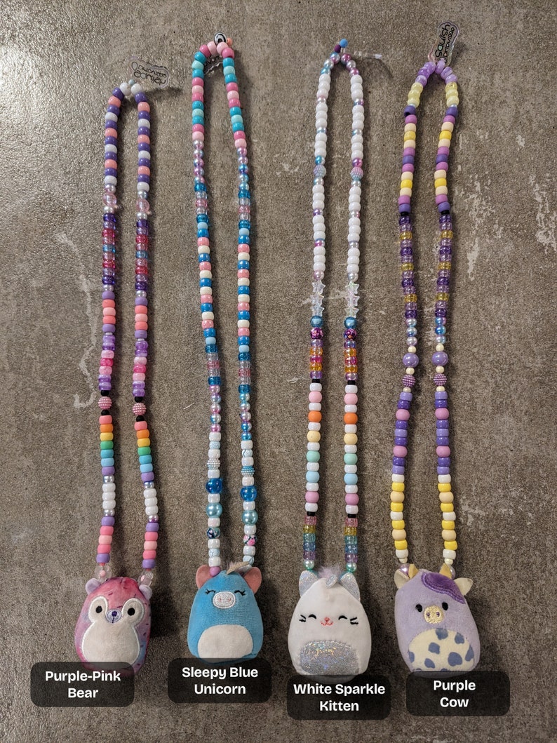 May include: Four beaded necklaces with plush animal pendants. The necklaces feature colorful beads in various sizes and shapes. The pendants include a purple-pink bear, a sleepy blue unicorn, a white sparkle kitten, and a purple cow. Each necklace has a tag with the name of the animal.