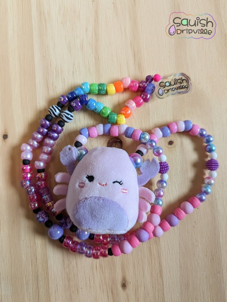 May include: A beaded necklace featuring a plush, lavender axolotl charm. The necklace has multiple strands of colorful beads, including rainbow, pink, purple, and black. The text "Squish Dripville" is visible.