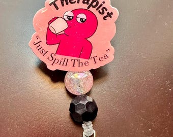 May include: A badge reel featuring a pink, die-cut acrylic topper with a cartoon character drinking from a mug. The text reads Therapist and Just Spill The Tea. Below the topper are two beads, one iridescent white and one faceted black.