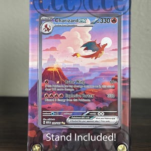 May include: A Charizard EX Pokémon card, stage 2, displayed in a protective case with a clear stand. The card features a red and orange dragon against a sunset landscape. The text "Stand Included!" is visible.