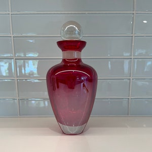 Może przedstawiać: Czerwona szklana butelka z przezroczystym szklanym korkiem. Butelka ma zaokrąglone dno i wąską szyjkę.