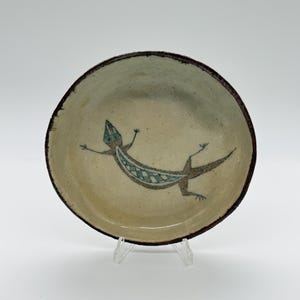 May include: A round, ceramic bowl with a hand-painted lizard design. The bowl has a beige interior with a brown rim. The lizard is depicted in shades of blue and brown. The bowl is displayed on a clear stand.
