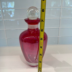 May include: A red glass bottle with a clear glass stopper. The bottle is approximately 7 inches tall.