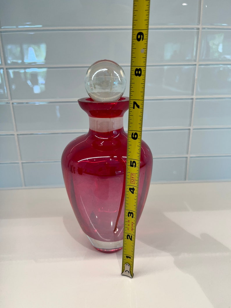 May include: A red glass bottle with a clear glass stopper. The bottle is approximately 7 inches tall.