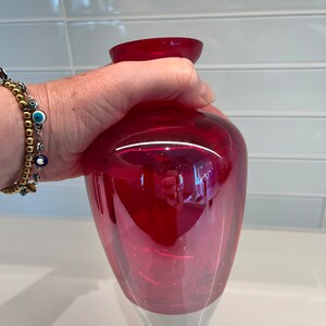 May include: A deep red glass vase with a rounded base and a slightly flared opening. The vase is clear and has a slight iridescent quality.