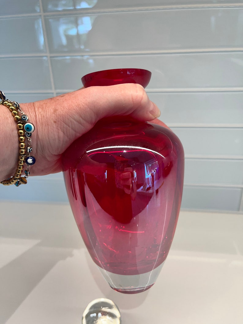 May include: A deep red glass vase with a rounded base and a slightly flared opening. The vase is clear and has a slight iridescent quality.