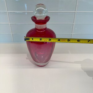 May include: A red glass bottle with a clear glass stopper. The bottle is approximately 6 inches tall.