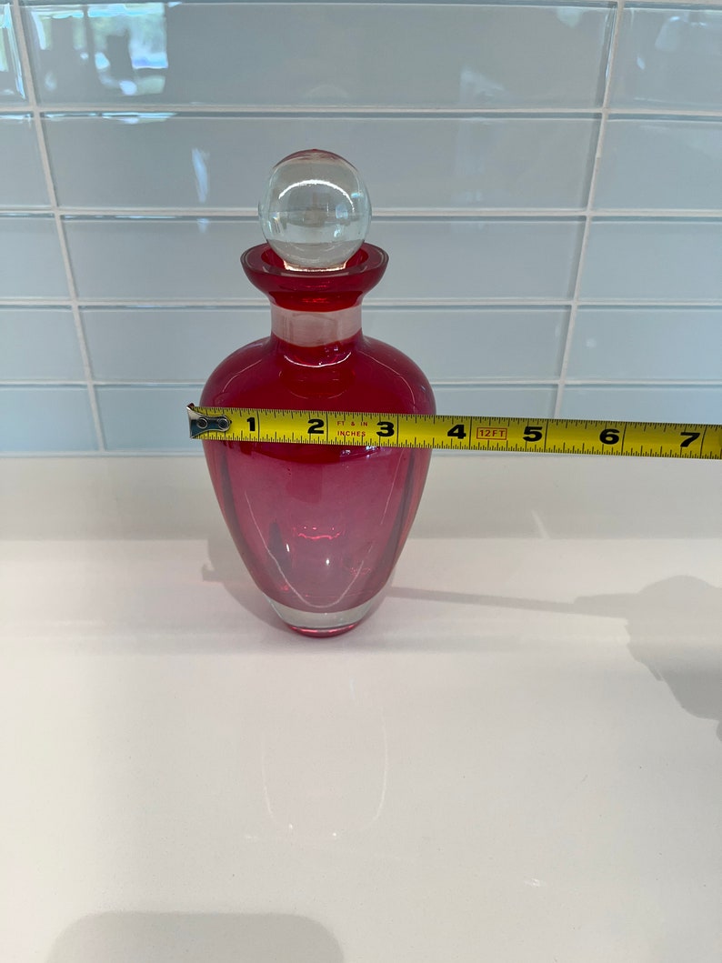 May include: A red glass bottle with a clear glass stopper. The bottle is approximately 6 inches tall.