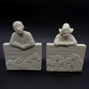 May include: A pair of off-white ceramic bookends. Each bookend features a figure leaning over a brick wall with tulip detailing. The figures are dressed in traditional Dutch attire, with a hat and a bonnet.