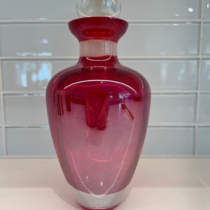 May include: A deep red glass bottle with a clear glass stopper. The bottle has a rounded bottom and a wide mouth.