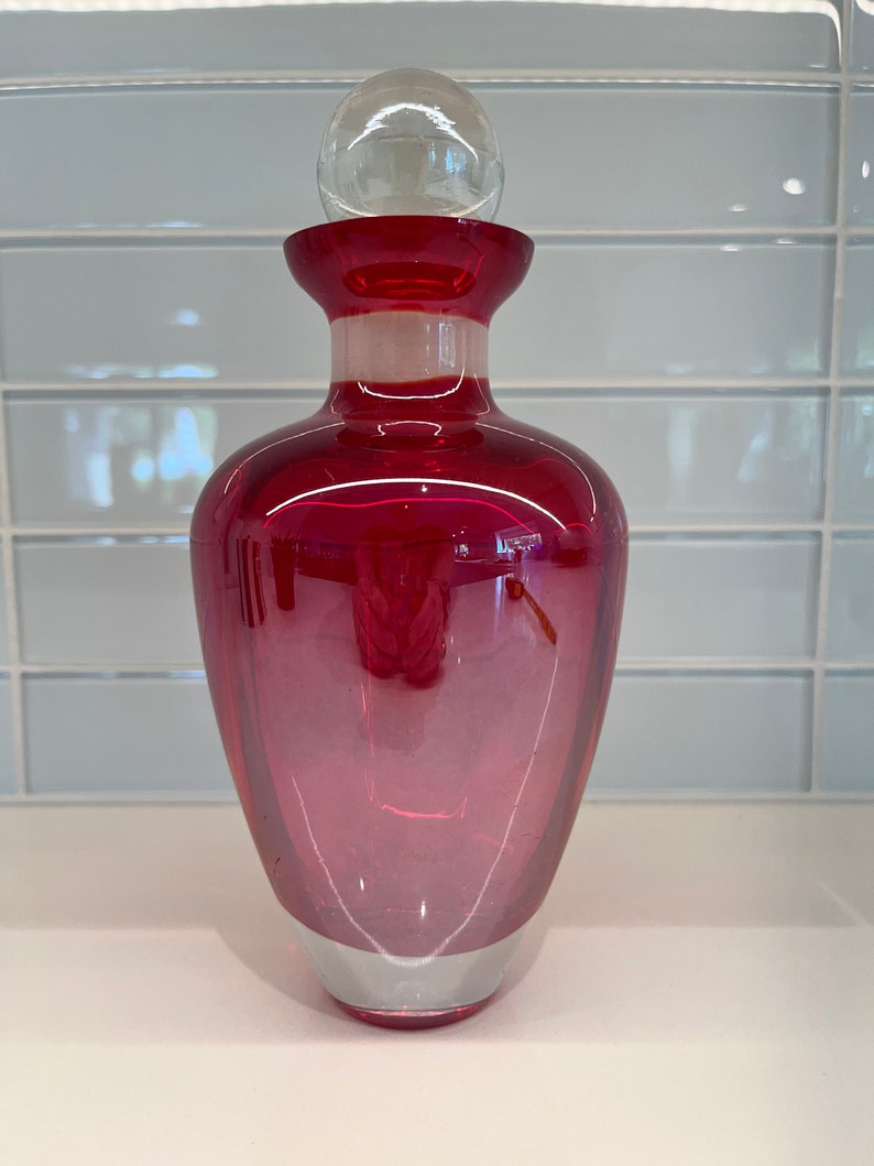 May include: A deep red glass bottle with a clear glass stopper. The bottle has a rounded bottom and a wide mouth.