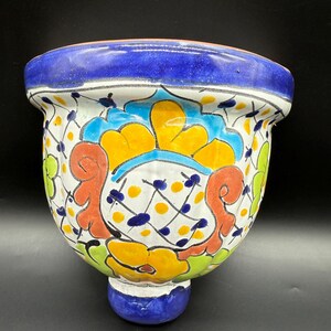 May include: A hand-painted ceramic wall planter with a vibrant floral design. The planter features a blue rim and base, with a white body adorned with yellow, orange, and green floral patterns. The planter is approximately 20 cm in diameter.