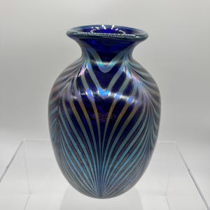 Könnte beinhalten: Eine kleine, kobaltblaue Glasvase mit einem irisierenden, federartigen Muster. Die Vase hat einen runden Körper und eine leicht ausgestellte Öffnung.