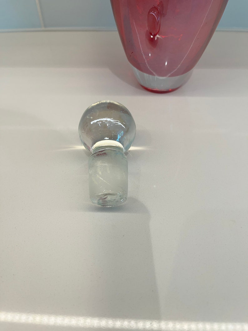 May include: A clear glass bottle stopper with a round top and a textured base. The stopper is designed to fit a bottle with a standard neck size.