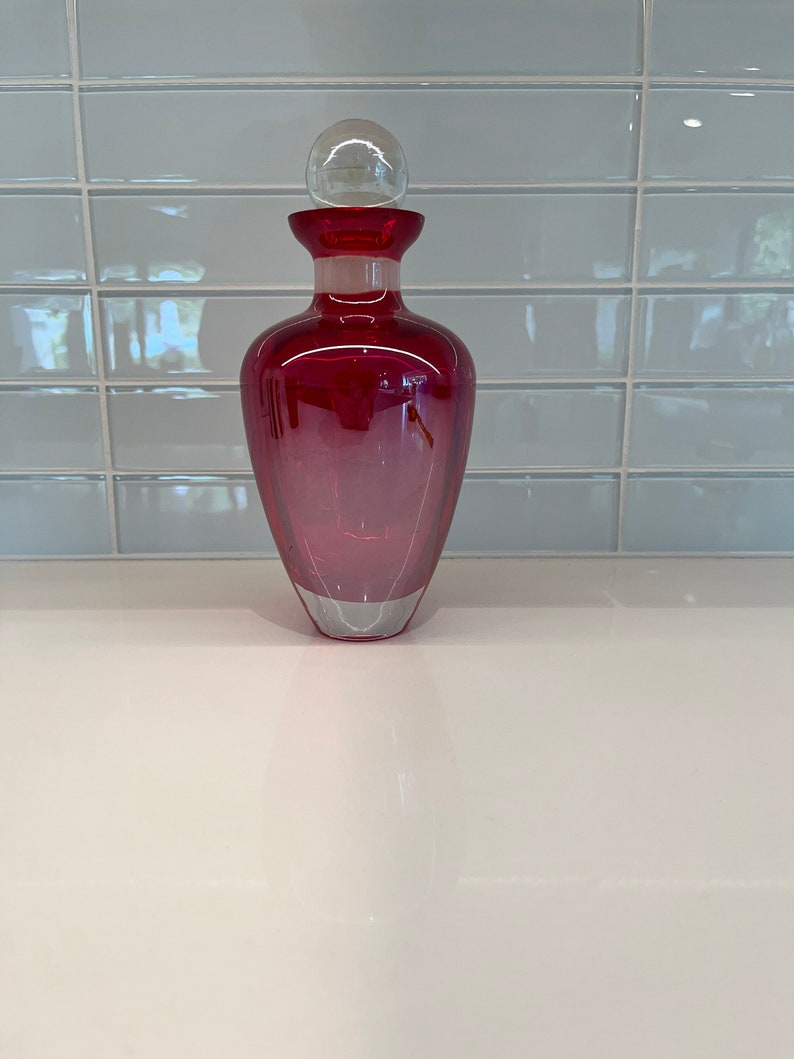 May include: A red glass bottle with a clear glass stopper. The bottle has a rounded base and a narrow neck.