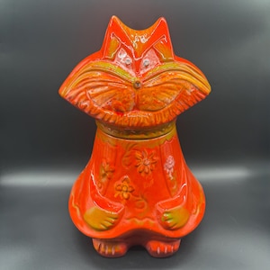 Pode incluir: Jarro de biscoitos de cerâmica laranja em forma de gato com um padrão floral e uma expressão caprichosa. O gato tem uma boca larga e aberta e olhos grandes e redondos.