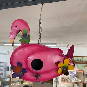 Könnte beinhalten: Ein pinkfarbenes Metall-Flamingo-Vogelhaus mit einem schwarzen Eingang und dekorativen Blumenakzenten. Das Vogelhaus ist an einer Kette aufgehängt und hat einen gelben Schnabel und bunte Blumendetails.