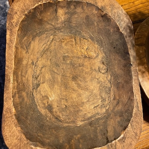 Könnte beinhalten: Eine große, rustikale Holzschale mit einem natürlichen braunen Finish. Die Schale ist oval geformt und hat einen tiefen, abgerundeten Innenraum. Sie hat eine raue, strukturierte Oberfläche und zeigt Anzeichen von Alter und Gebrauch.