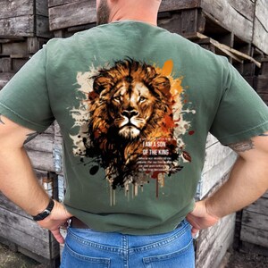 May include: Olive green t-shirt featuring a detailed lion's head graphic with orange, brown, and white accents. The text "I AM A SON OF THE KING" is printed below the lion's head. The shirt is made of a soft material.