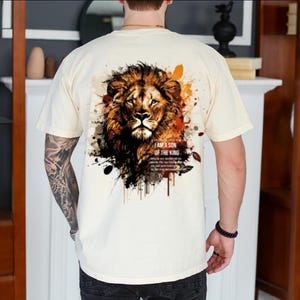 May include: Cream-colored t-shirt featuring a detailed lion's head graphic on the back, with text that reads "I AM A SON OF THE KING." The lion design incorporates shades of brown, orange, and black, with paint splatter effects.