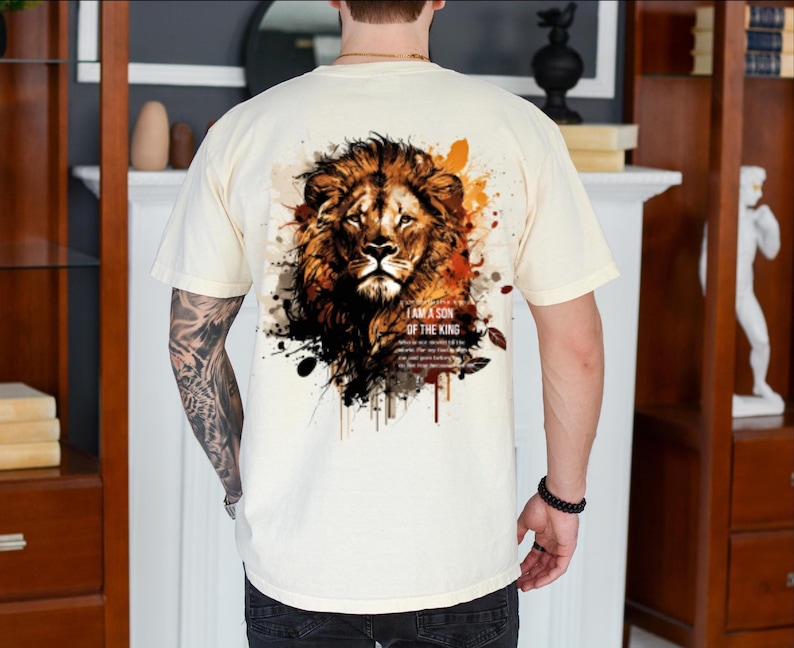 May include: Cream-colored t-shirt featuring a detailed lion's head graphic on the back, with text that reads "I AM A SON OF THE KING." The lion design incorporates shades of brown, orange, and black, with paint splatter effects.