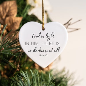 Puede incluir: Adorno blanco en forma de corazón con la inscripción "God is light, in Him there is no darkness at all" y la referencia "1 John 1:5". Cuelga de un hilo dorado, sobre un fondo de follaje verde y dorado.