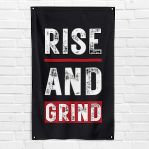 Puede incluir: Bandera negra con las palabras "RISE AND GRIND" en fuente blanca desgastada. Las palabras están separadas por una línea roja y un cuadro rojo. La bandera cuelga contra una pared de ladrillos blancos.