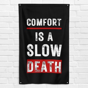 Puede incluir: Bandera de pared negra con el texto "COMFORT IS A SLOW DEATH" en blanco, con una línea y un recuadro rojos. La bandera es rectangular y cuelga de dos ojales en la parte superior. El texto tiene un aspecto envejecido.