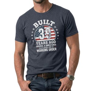 May include: A navy blue t-shirt with a graphic that reads "BUILT 39 YEARS AGO" in white text, with an American flag design. The shirt also includes the text "ORIGINAL & UNRESTORED most parts still in WORKING ORDER".