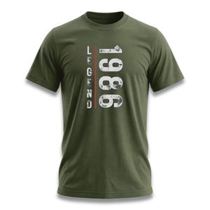 以下が含まれることがあります： オリーブグリーンのTシャツ。左側に縦に重ねられた「LEGEND」の文字、右側に白の掠れたフォントで「1986」の数字がプリントされています。テキストの中央には赤い縦線が入っています。